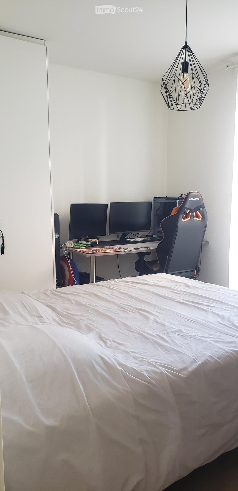 A modern bedroom with a large bed covered in white sheets, a gaming chair, a desk with multiple screens, and a hanging geometric light fixture.