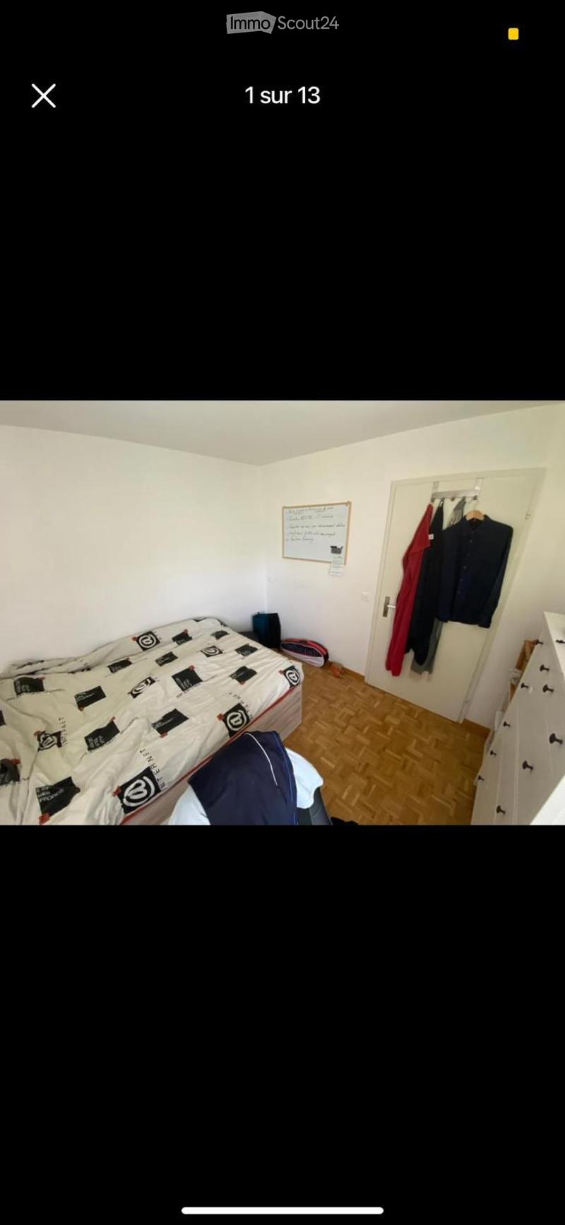 A small bedroom with a bed, a wardrobe, and a chair. The room features wooden flooring and white walls.