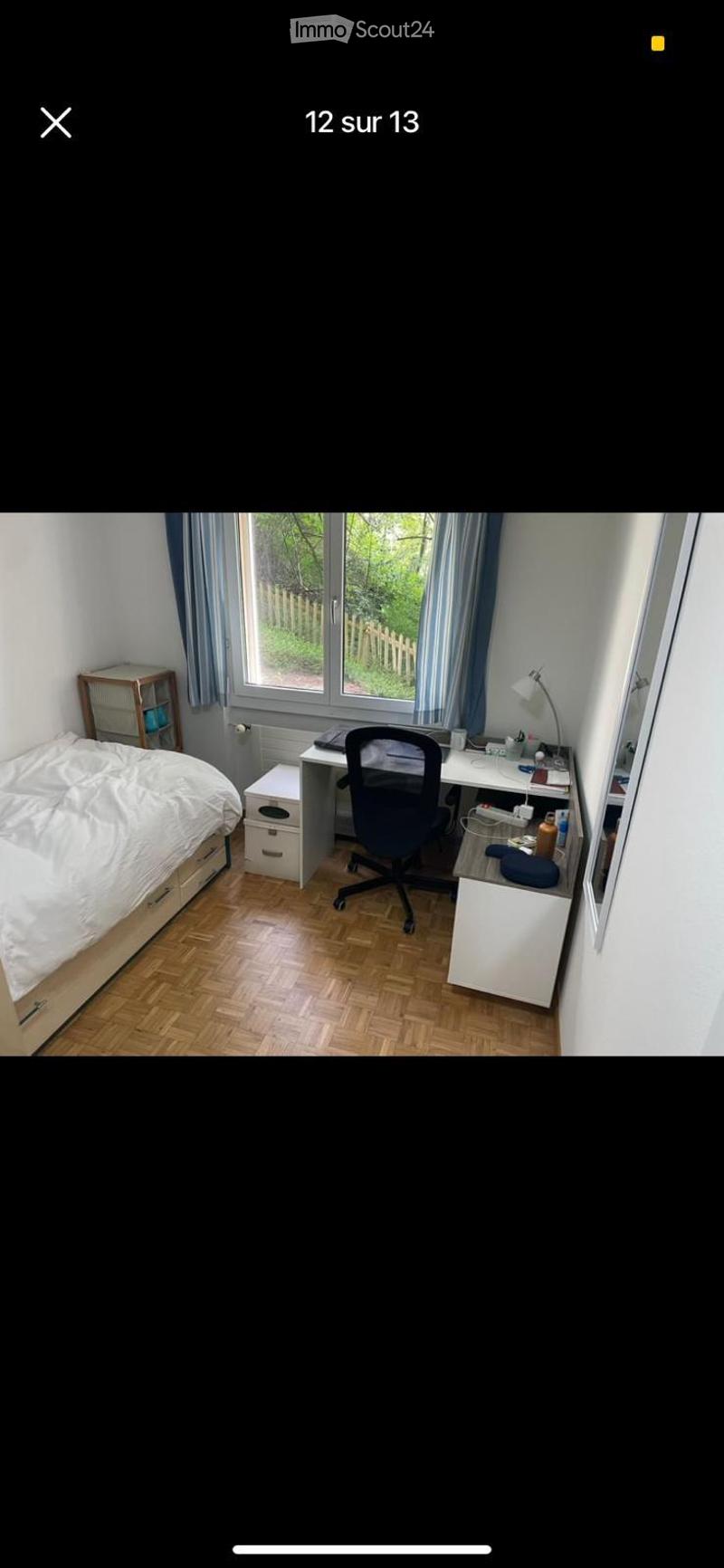 Cozy bedroom with a wooden floor, large window with blue curtains, a bed with white bedding, a desk with a chair, a lamp, and a small storage unit.
