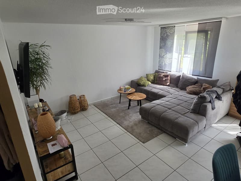 Modern living room with a grey sectional sofa, coffee table, side tables, and decorative vases. The room features tiled flooring, large windows with curtains, and a television mounted on the wall.