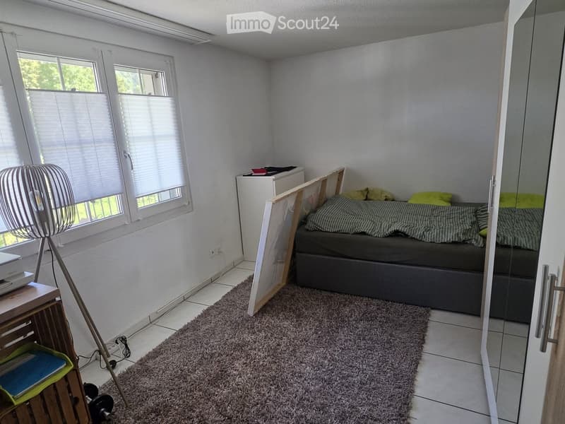 Cozy bedroom with a bed, fan, small table, and mirror. The room features a large window with blinds and a tiled floor.