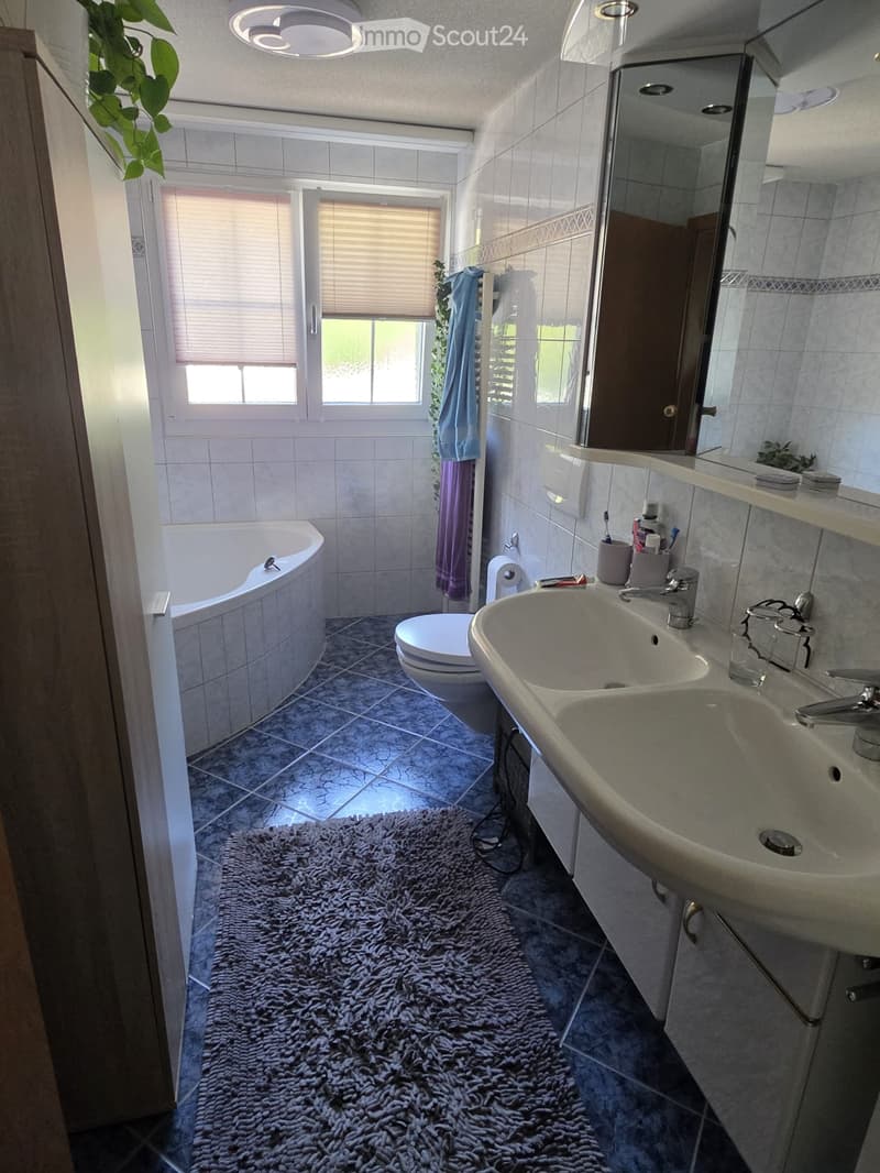 Bathroom with a bathtub, toilet, double sink, mirror, and a fluffy rug on the floor. The room has tiled walls and a window with blinds.