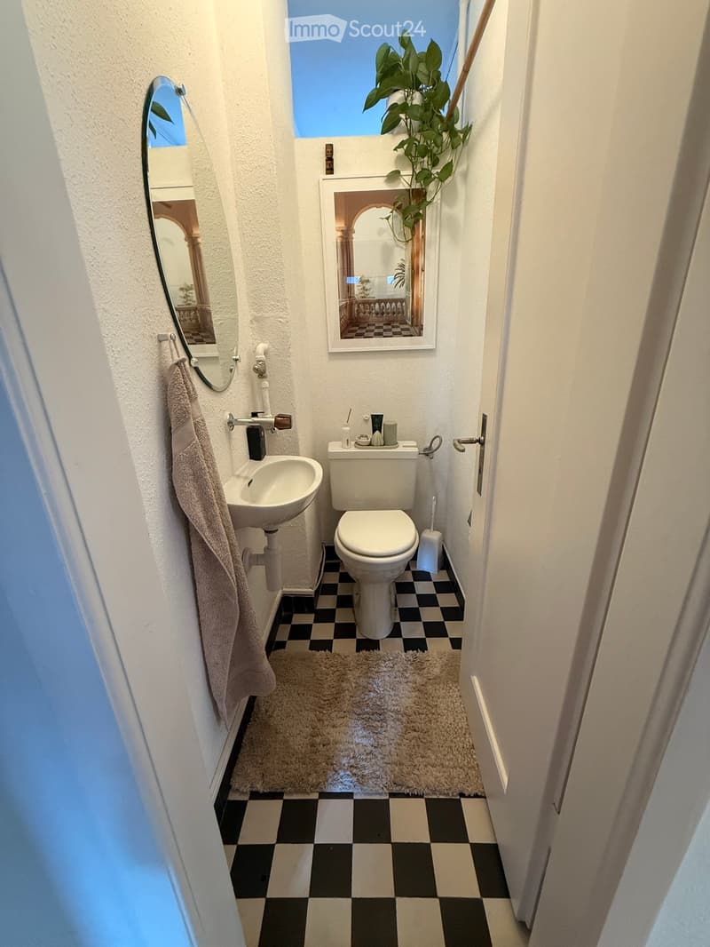Compact bathroom with white walls, black and white checkered floor, a toilet, sink, mirror, towel, and a small plant on the windowsill.