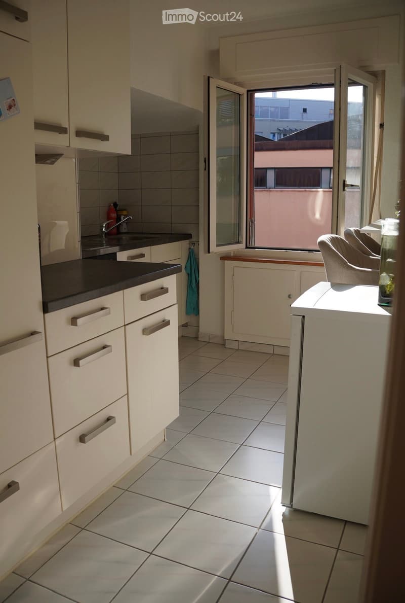 Modern kitchen with white cabinets, black countertop, tiled floor, and a small seating area with a chair and table near a large window.