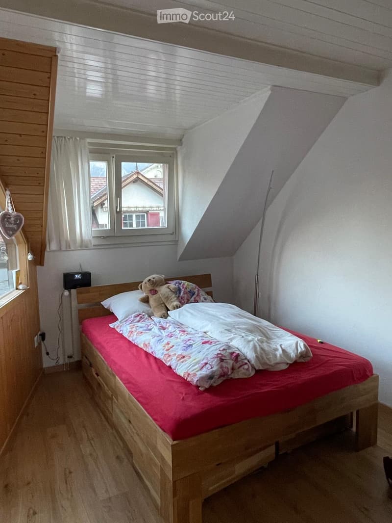 Cozy attic bedroom with wooden bed, red bedding, floral duvet, teddy bear, and a window overlooking a house.