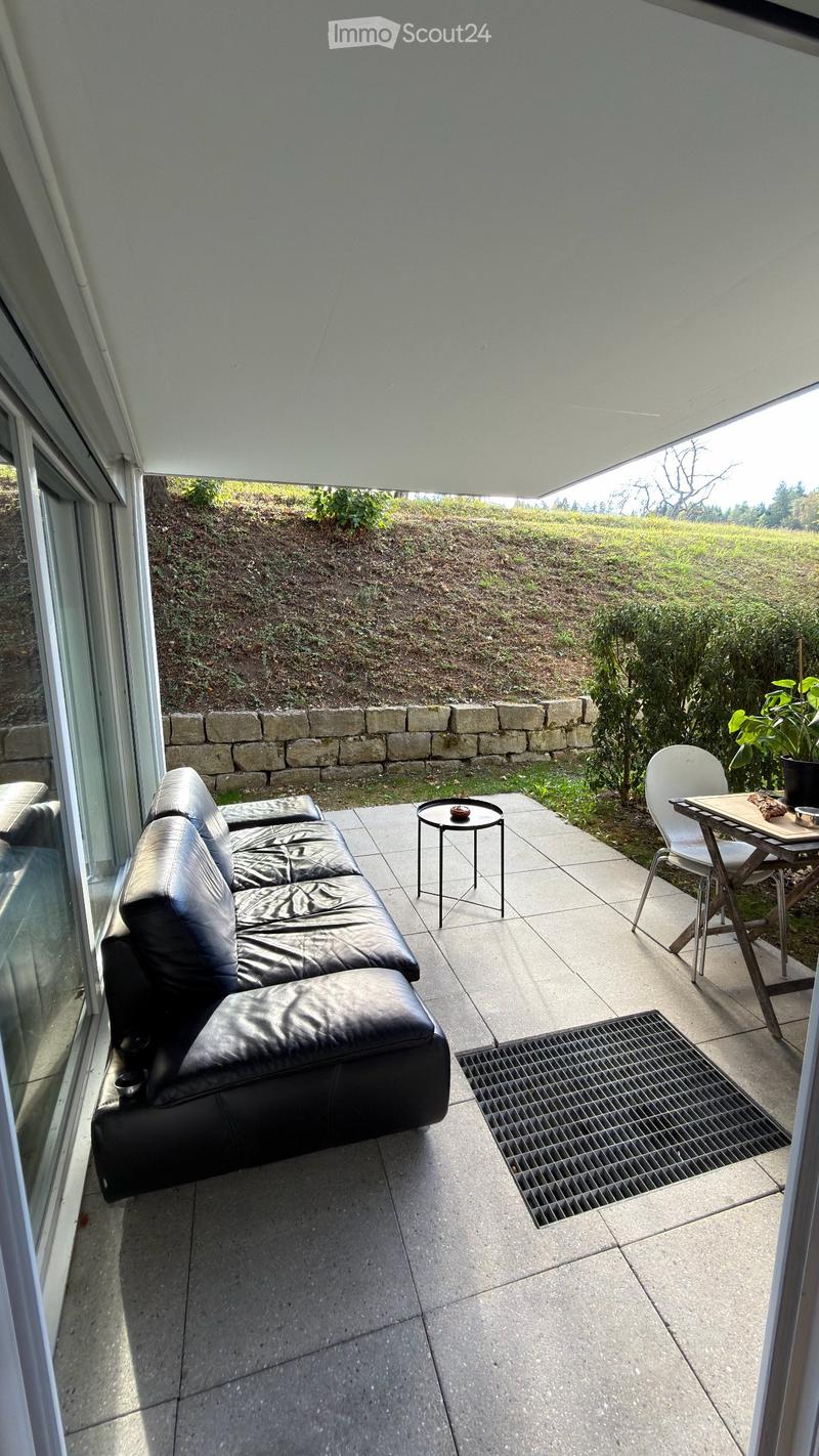 Modern balcony with a black leather sofa, small round table, folding chair, and potted plant, overlooking a landscaped garden.
