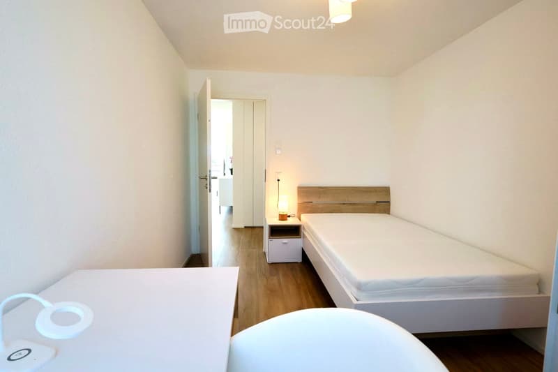 A small, neatly furnished bedroom with a single bed, a nightstand with a lamp, and a drawer unit. The room has light wooden flooring and white walls.