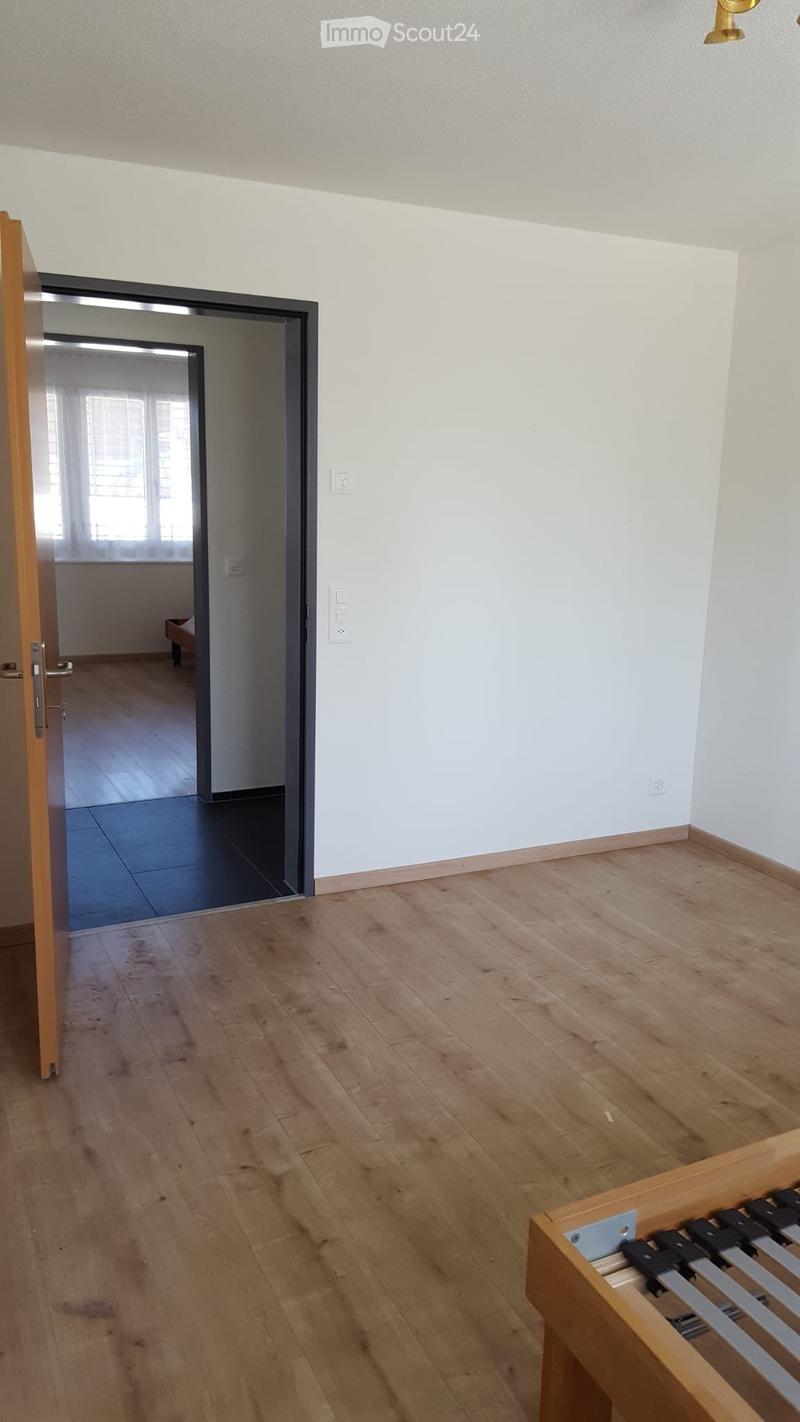 Empty room with wooden flooring, white walls, and an open doorway leading to another room with tiled flooring and a window.