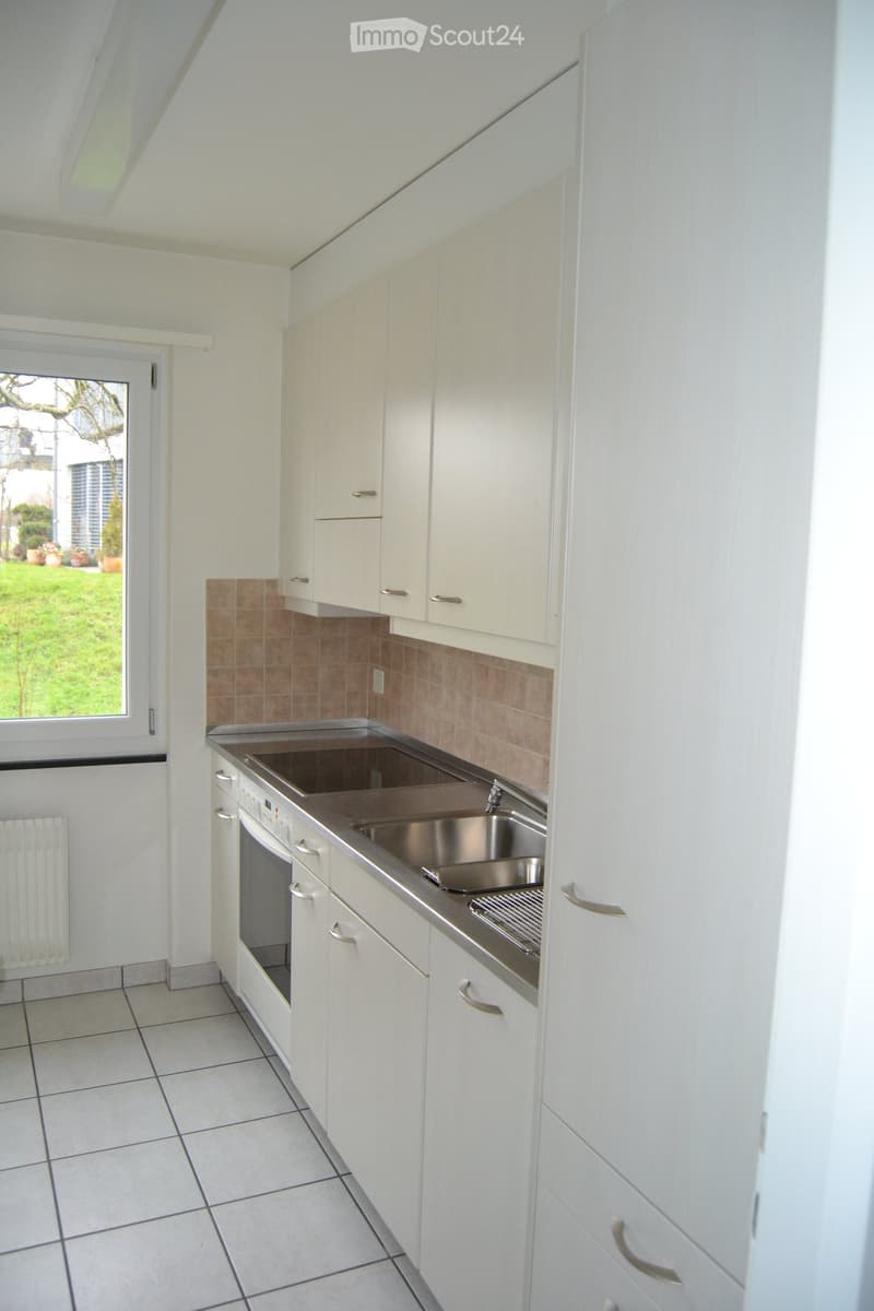 Modern kitchen with white cabinets, stainless steel sink, and tiled floor. Features a window with a view of a garden.