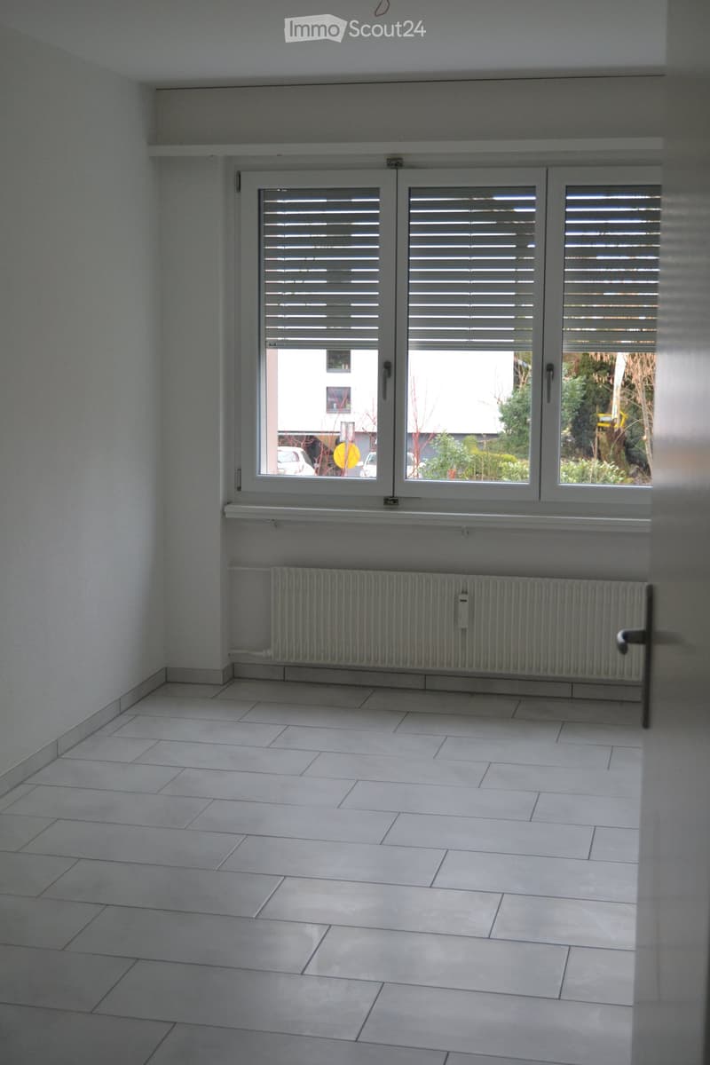 Empty room with white tiled flooring, white walls, large window with blinds, radiator, and a door handle visible.