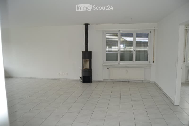 Empty room with tiled floor, white walls, large window with blinds, and a black wood-burning stove with chimney.