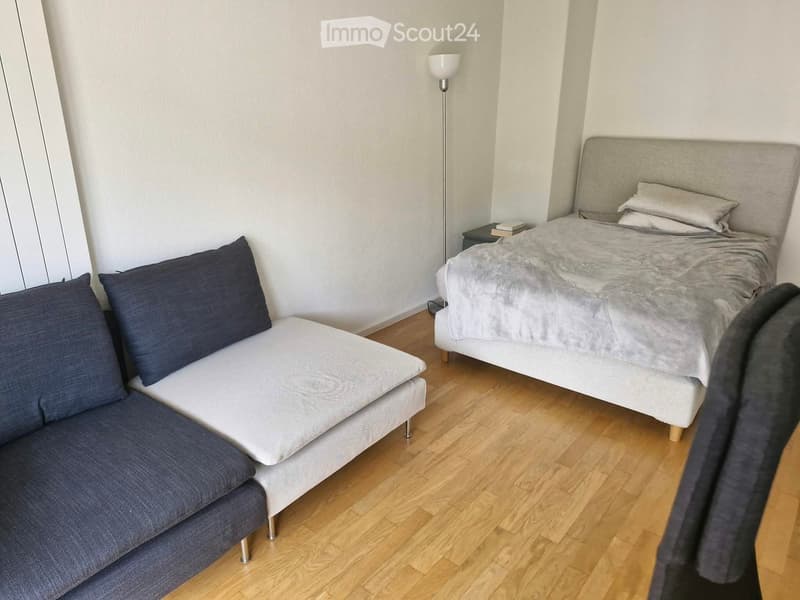 A modern, compact bedroom with a double bed, a small sofa, a nightstand, and a floor lamp. The room features light wooden flooring and neutral-colored walls.