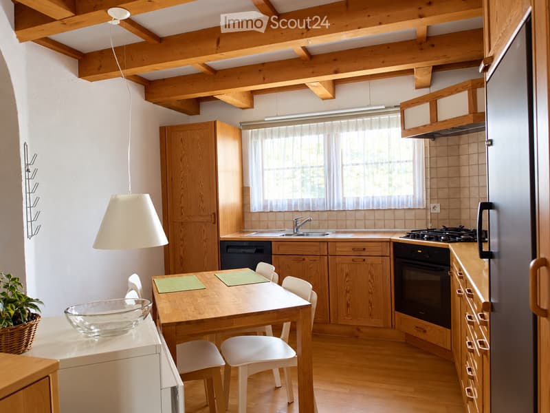 Bright kitchen with wooden cabinets, a central dining table with chairs, a refrigerator, oven, sink, and a hanging lamp.