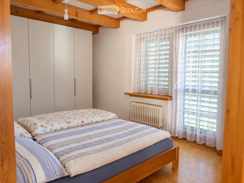 Cozy bedroom with wooden bed, floral and striped bedding, white wardrobe, wooden shelf, radiator, and sheer curtains on windows.