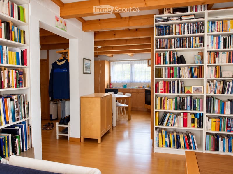A cozy, well-lit room with wooden floors and exposed wooden beams. Features include bookshelves filled with books, a small dining area with a table and chairs, a coat hanging on a rack, and a cabinet. The room has a warm, inviting atmosphere with natural