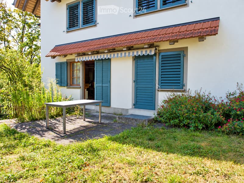 A small outdoor patio area with a metal table, surrounded by a well-maintained lawn and colorful flowering bushes. The house features a red-tiled roof, white walls, and blue shutters.