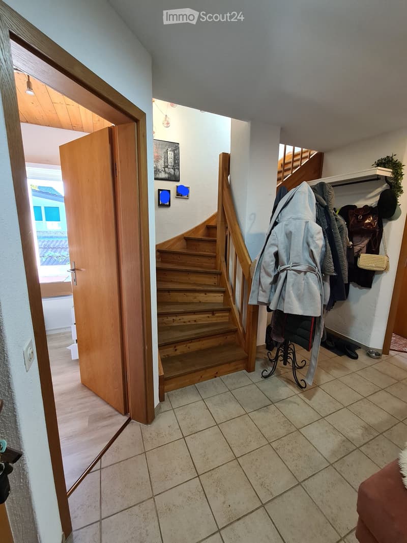 Hallway with tiled floor, wooden staircase, coat rack with jackets and bags, and an open door leading to another room.