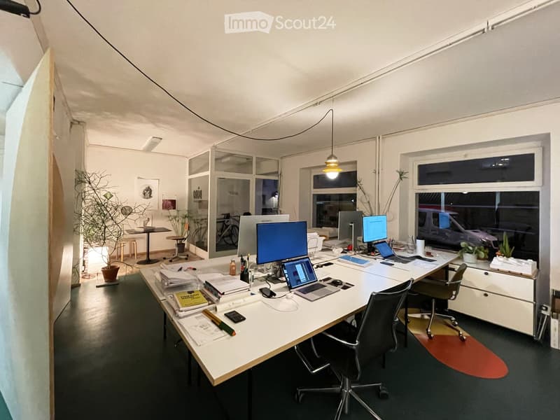 Modern office space with a large desk, multiple computer screens, laptops, office chairs, and a small meeting area with a table and stools. The room features large windows, pendant lighting, and decorative plants.