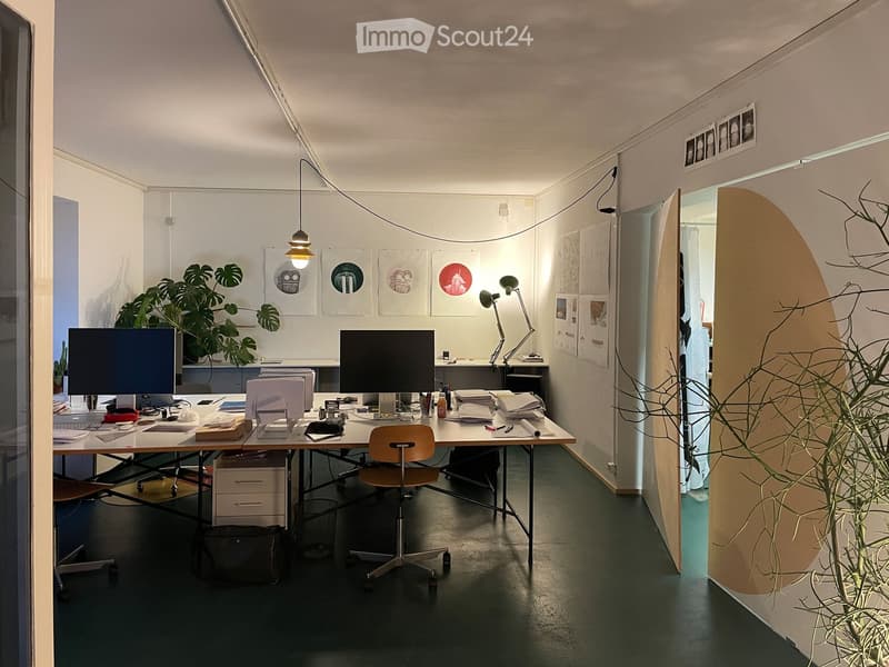 Modern office space with a large desk, two computer monitors, a chair, a lamp, and several framed artworks on the wall. There is a plant in the corner and a cabinet with drawers.