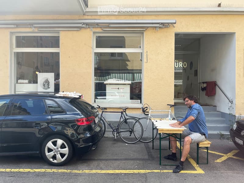 A man sits at a small table outside a building with large windows and a sign reading "URO." A bicycle and a car are parked nearby.