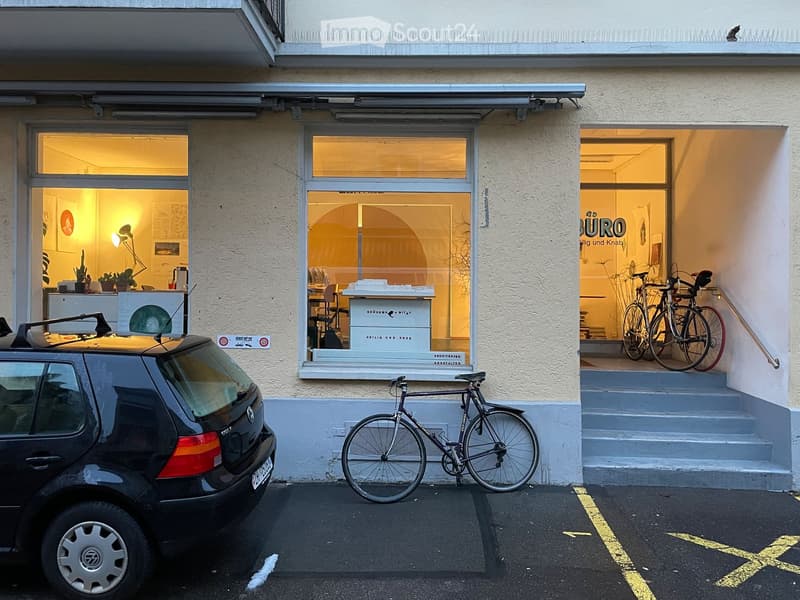 Facade of a building with large windows and a storefront, bicycles parked outside, and a car in the parking area.