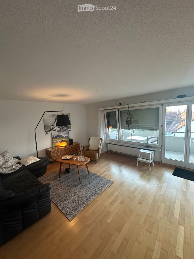 Modern living room with wooden flooring, large windows, and a balcony access. Features a black sectional sofa, coffee table, armchair, side table, floor lamp, and a picture frame on the wall.