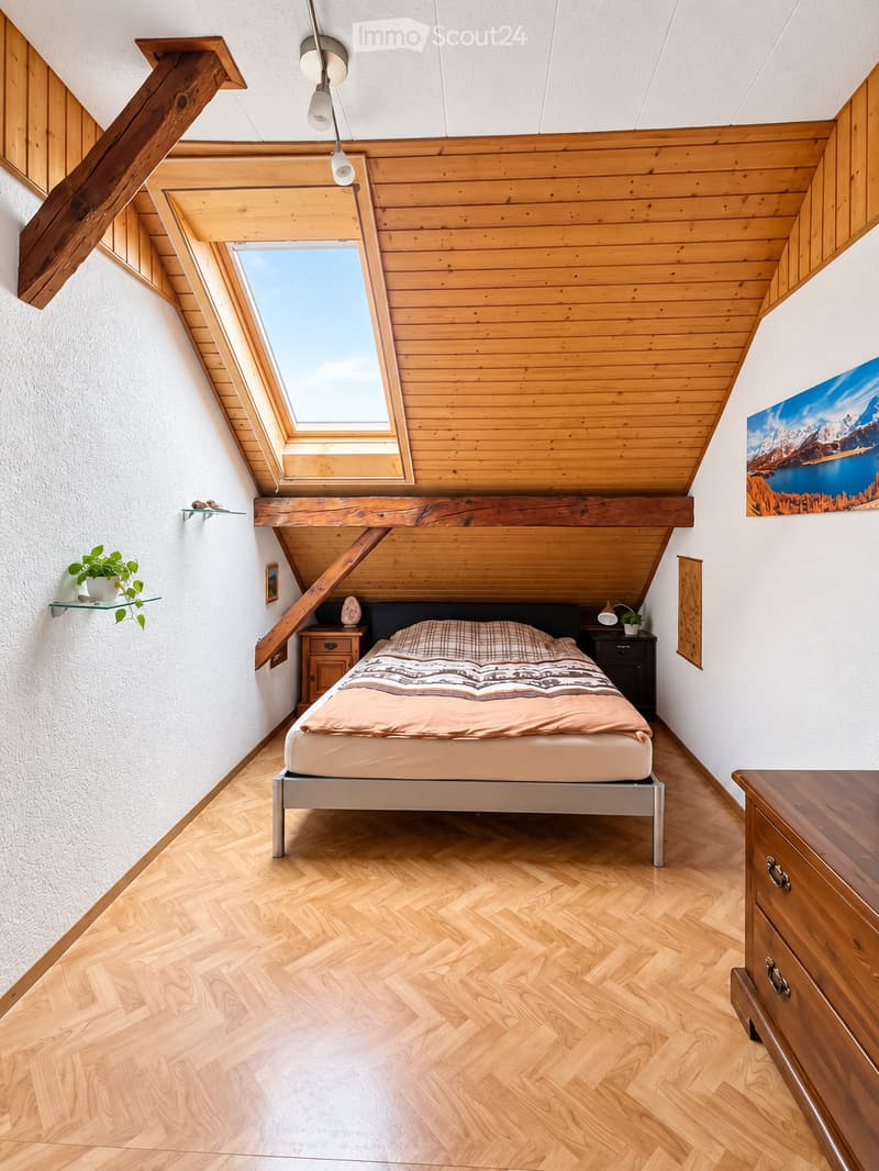 Cozy attic bedroom with wooden walls and herringbone flooring, featuring a bed, nightstands, a lamp, and a large skylight window.