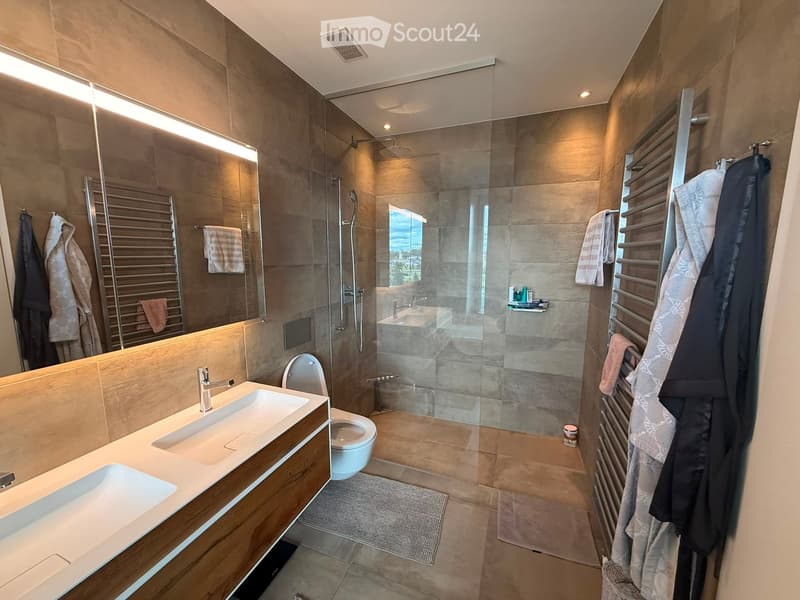 Modern bathroom with double sink, toilet, large mirror, glass-enclosed shower, towel racks, and towels.