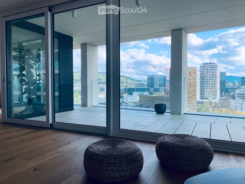 Modern living room with wooden flooring, large glass doors leading to a balcony with city view, two woven stools, and a small table.