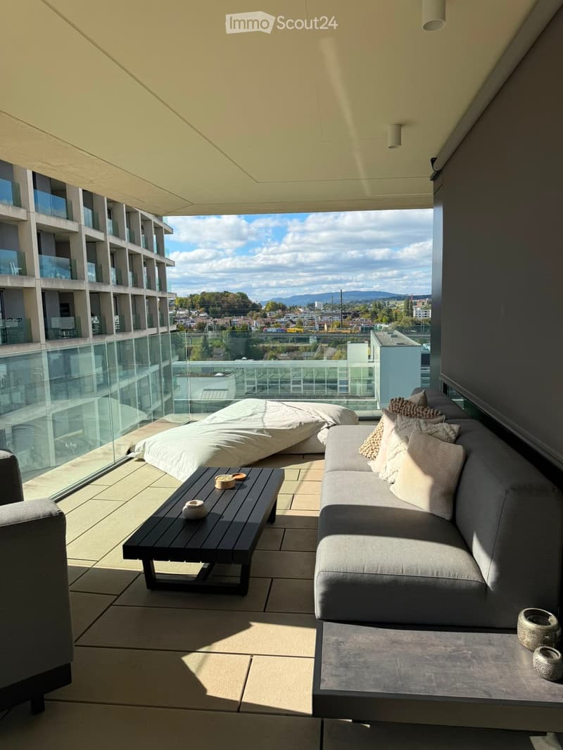 Modern balcony with a sofa, coffee table, bed, and decorative items, offering a scenic city view.