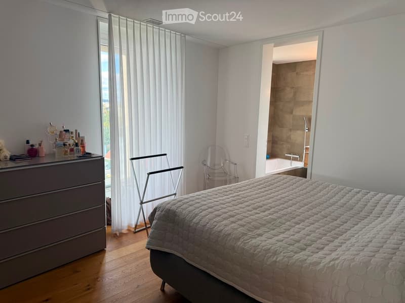 A modern bedroom with a large bed, a dresser with various items, a chair, and an open bathroom with a bathtub and shower.