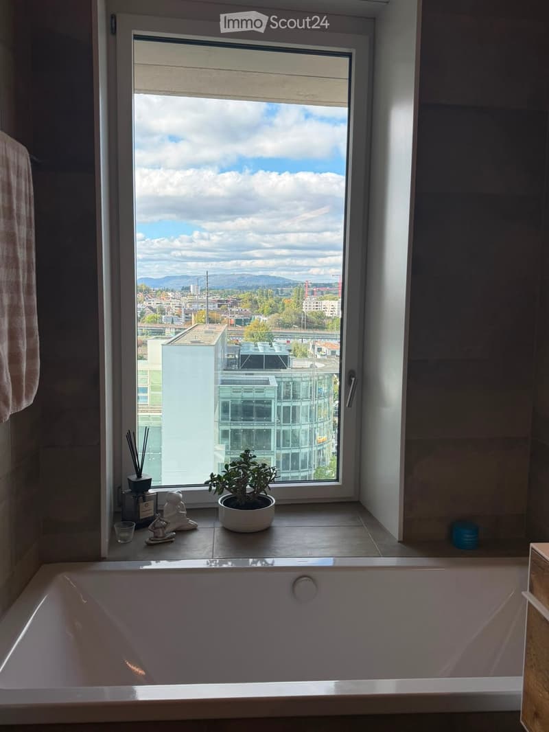 Bathroom with a large window offering a cityscape view, featuring a bathtub, a potted plant on the windowsill, an incense holder, a small cup, and a decorative statue.