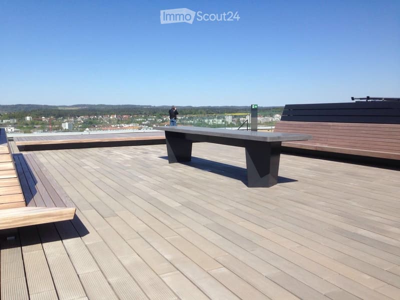 Modern rooftop terrace with wooden decking, a large rectangular table, and seating areas, offering a scenic city view.