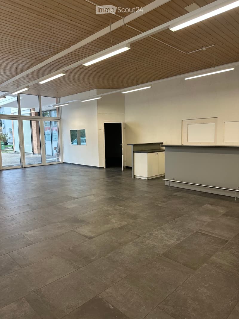 Empty room with tiled floor, white walls, and a reception desk area. Large windows and doors lead outside.