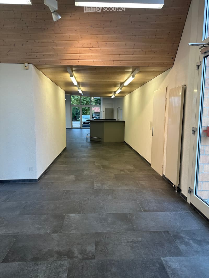 Empty hallway with tiled flooring, white walls, wooden ceiling, and recessed lighting. Features a reception desk and a glass door leading outside.