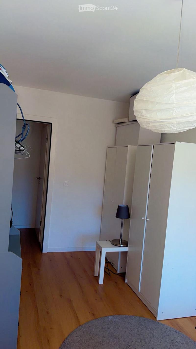A small, unfurnished room with light wooden flooring, white walls, a small table with a lamp, and a wardrobe. The room has a grey rug on the floor and a hanging paper lantern light fixture.
