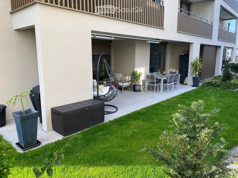 Well-maintained garden with a neatly trimmed lawn, a wicker storage box, a hanging wicker chair, a small seating area with a sofa and table, and several potted plants.