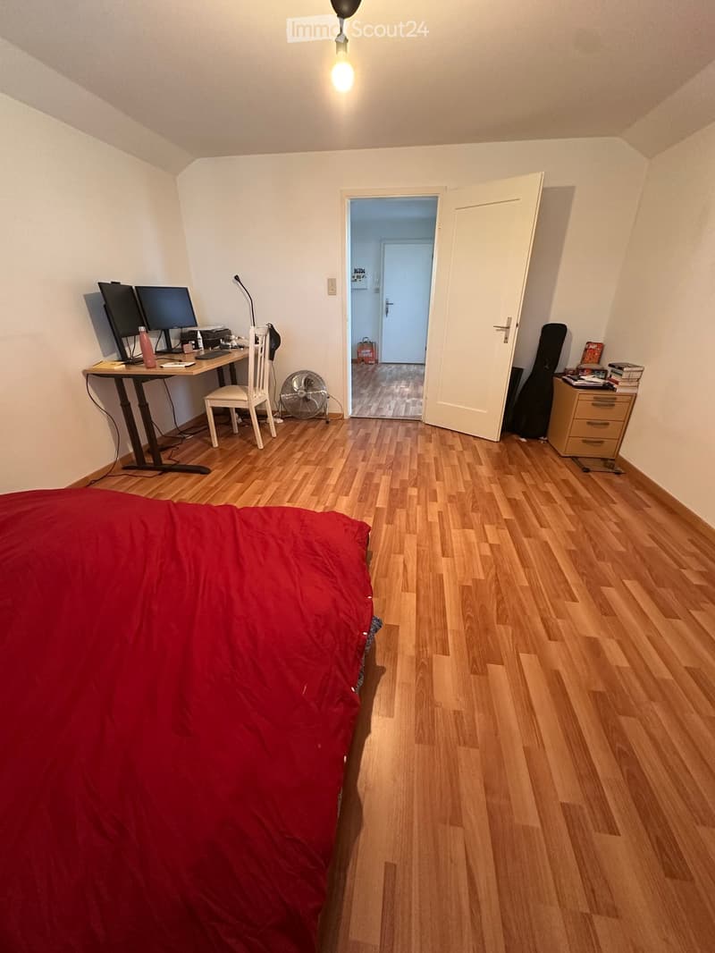 A furnished bedroom with a wooden floor, a bed with a red blanket, a desk with two screens, a chair, a fan, a guitar case, and a small drawer unit.