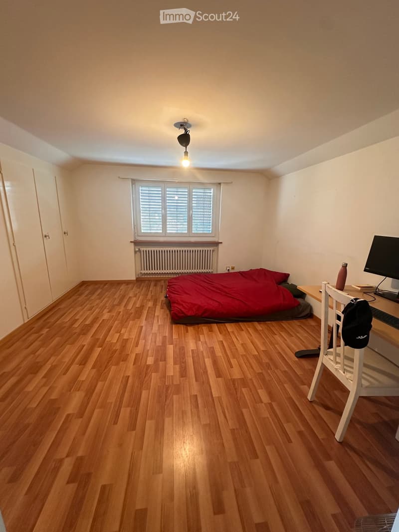 A small, furnished bedroom with wooden flooring, a bed with red bedding, a desk with a chair, a screen, and a backpack. The room has a window with blinds and a radiator below it.