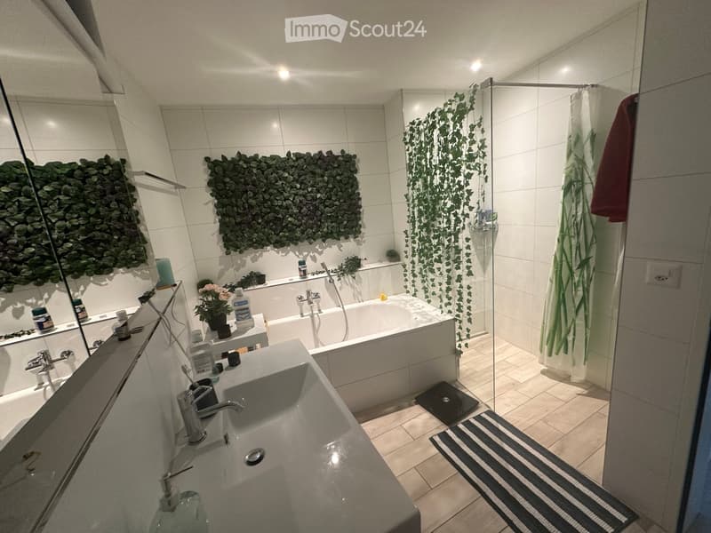 Modern bathroom with a white tiled design, featuring a sink, bathtub, and a glass-enclosed shower area with a leafy green curtain. The space includes a mirror, a towel, and decorative elements like a small plant and a candle on the sink counter.