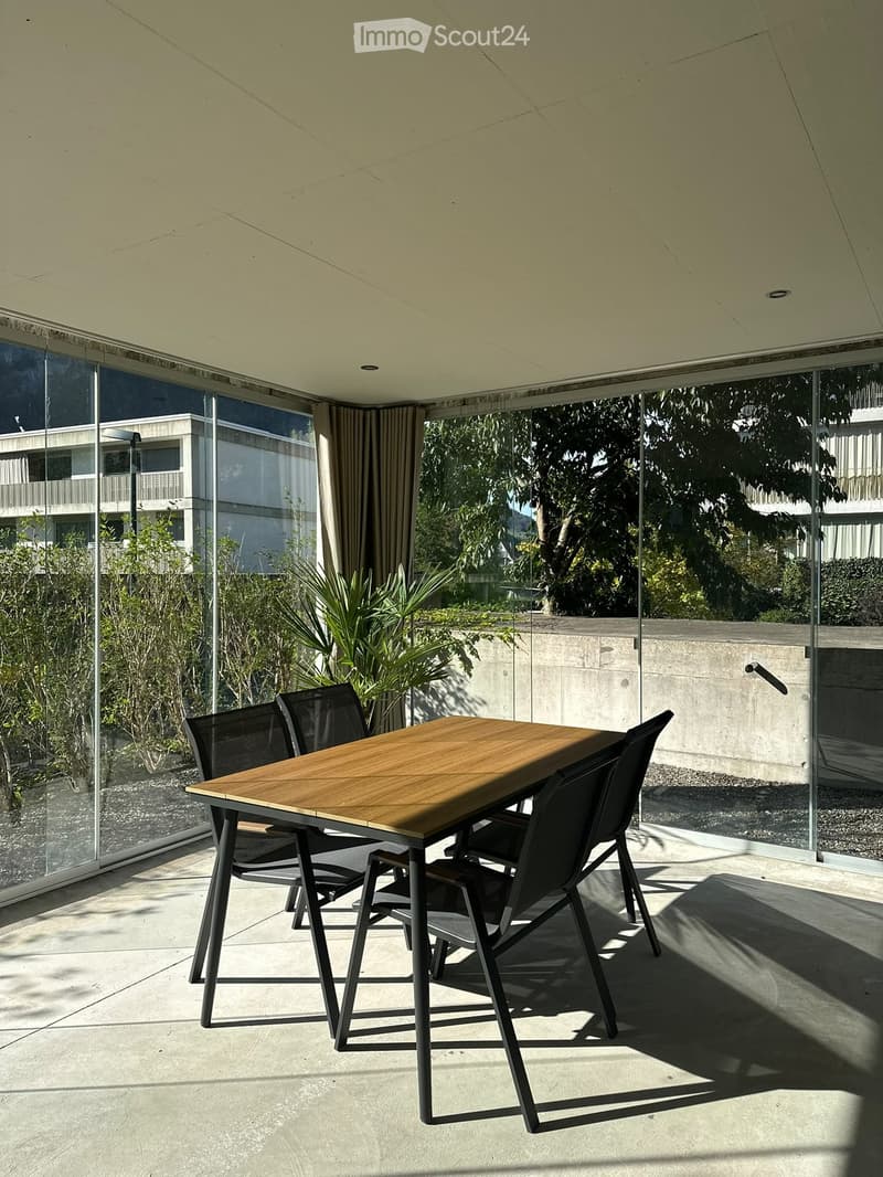 Modern outdoor dining area with a wooden table and four black chairs, large glass walls, and a view of greenery and buildings.