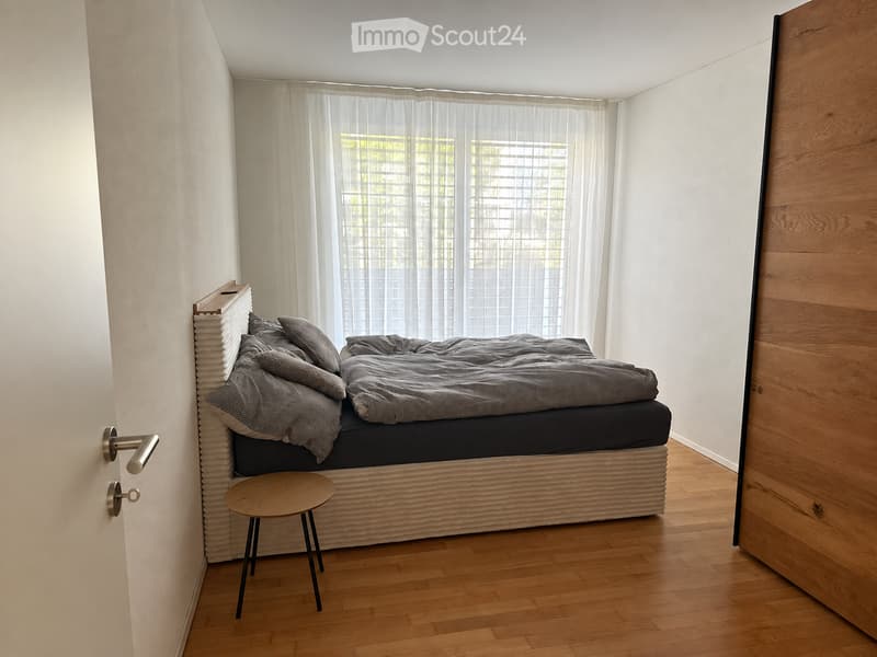 Modern bedroom with a wooden bed frame, grey bedding, a small wooden stool, and large windows with sheer curtains.
