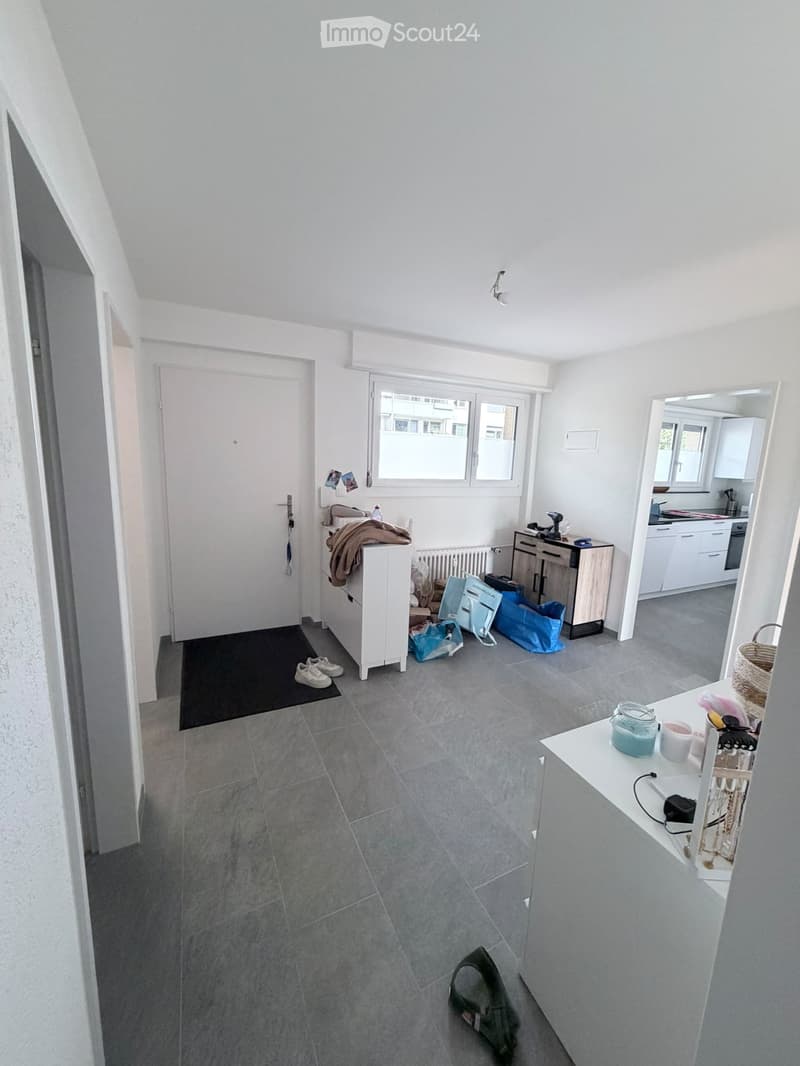 A modern, well-lit hallway with grey tiled flooring, white walls, and a large window. Visible items include a white cabinet, a mirror, a small table with a cup and bottle, a stool, a radiator, a door, and a window.
