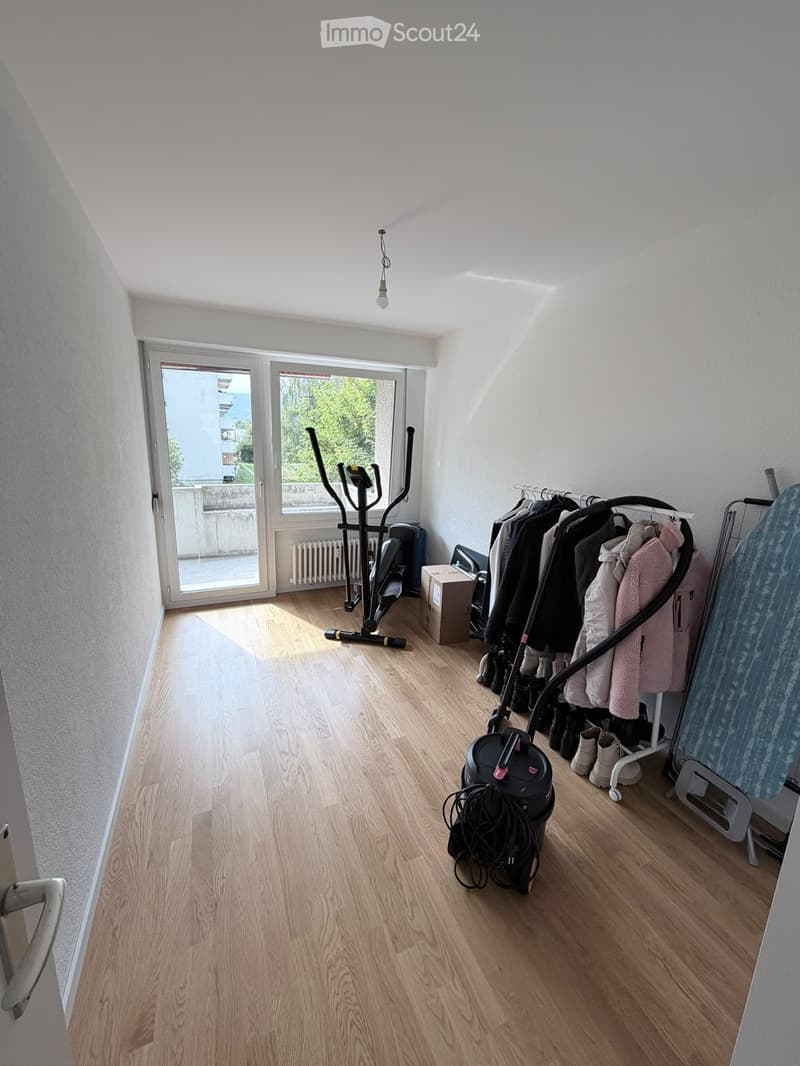 A well-lit room with wooden flooring, featuring an exercise bike, a clothing rack with coats and shoes, a vacuum cleaner, an ironing board, and a radiator near the window.