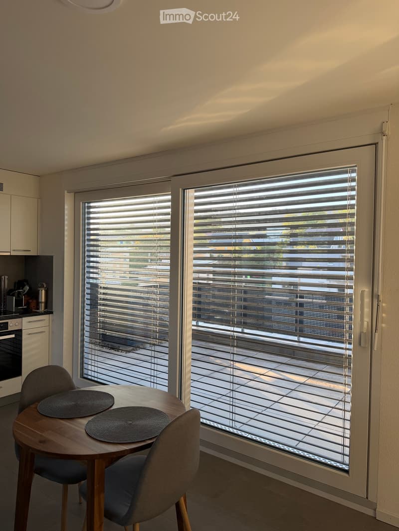 A modern dining area with a round wooden table, two chairs, and a view of a balcony through large windows with blinds. The kitchen area includes an oven and a small cabinet.