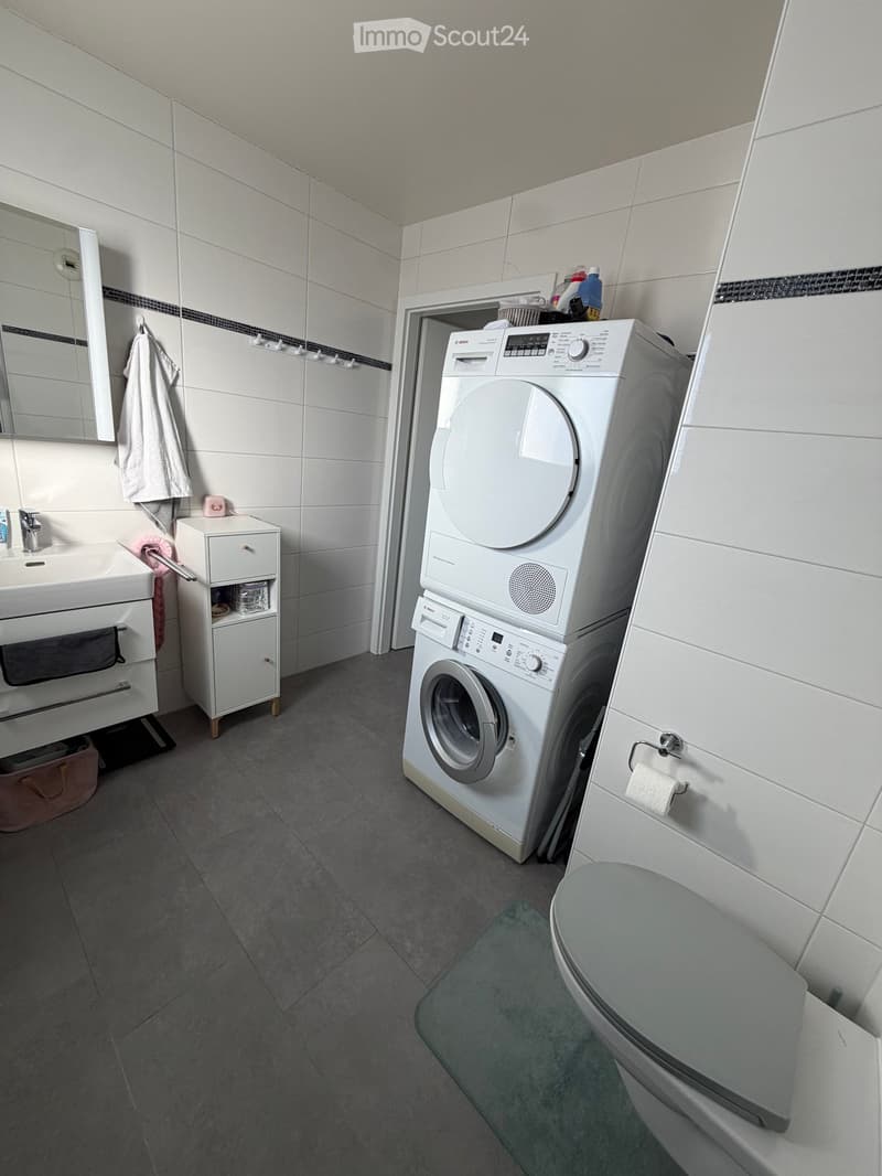 Modern bathroom with white tiles, a sink, mirror, cabinet, washing machine, and toilet. Includes a towel and toilet paper holder.