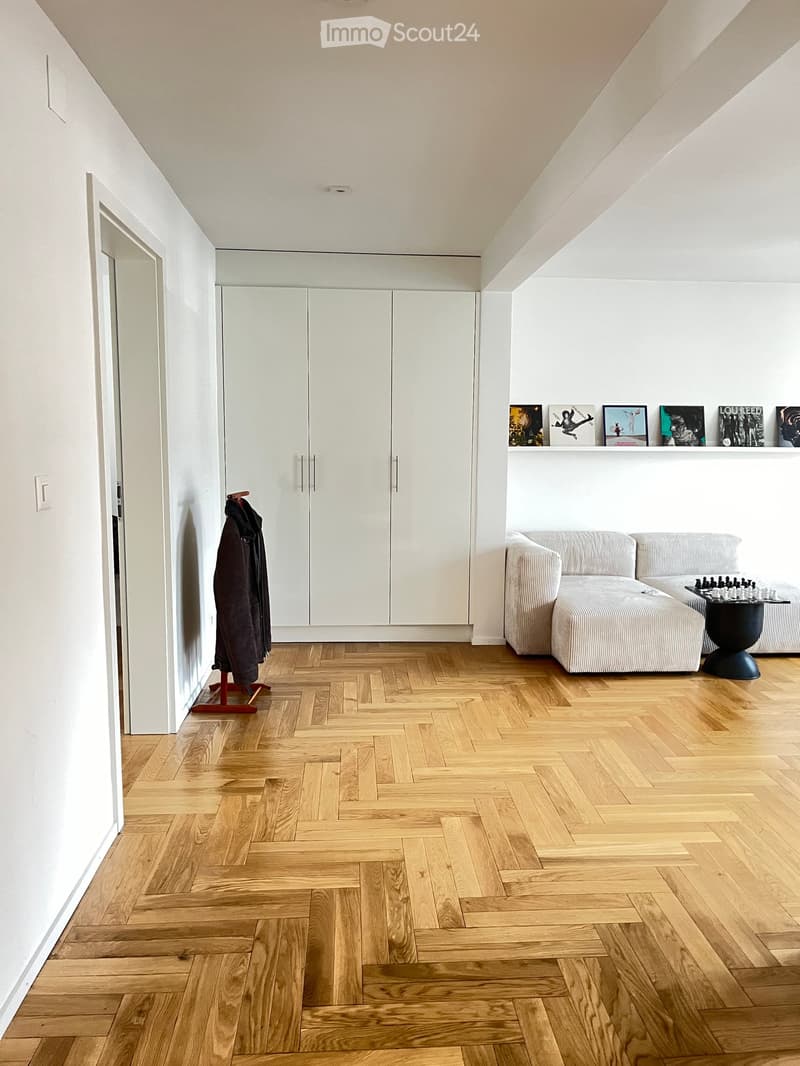 Modern living room with herringbone wooden flooring, white walls, a grey sectional sofa, a small black table with a chessboard, a coat rack with a jacket, and built-in white cabinets with framed artwork on the wall.