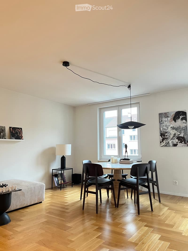 Bright, minimalist room with wooden herringbone flooring, large window, modern furniture including a sofa, dining table with chairs, and decorative items.