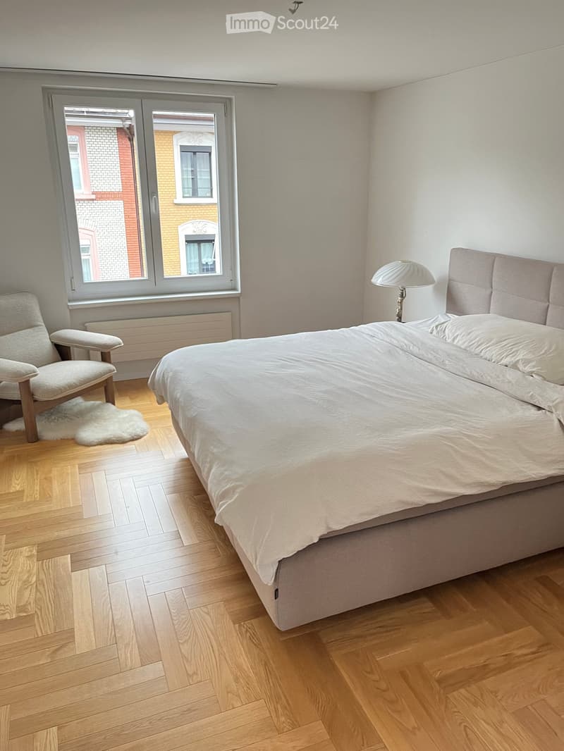Bright bedroom with wooden flooring, large window, bed with white bedding, armchair, and a lamp on the nightstand.