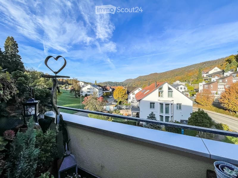 Balcony with metal railing, overlooking a residential area with houses, trees, and a clear blue sky.